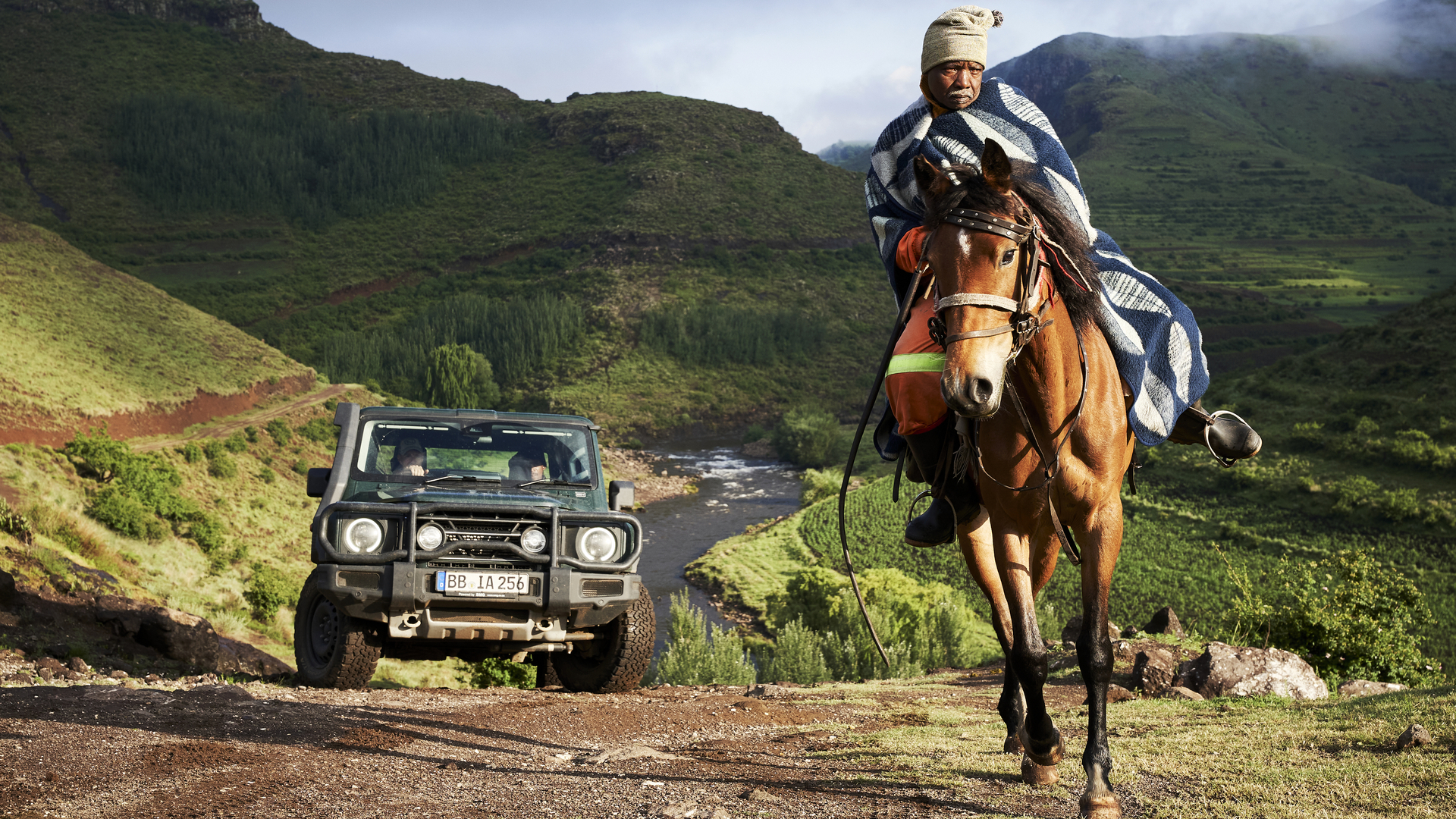 A man on horseback wearing traditional Basotho attire rides past an INEOS Grenadier Quartermaster navigating a rugged mountain trail in Lesotho.